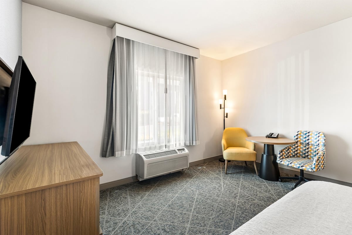 A hotel room with a wooden desk, a small round table, a patterned armchair, a bed, a large window with sheer curtains, and a wall-mounted air conditioner.