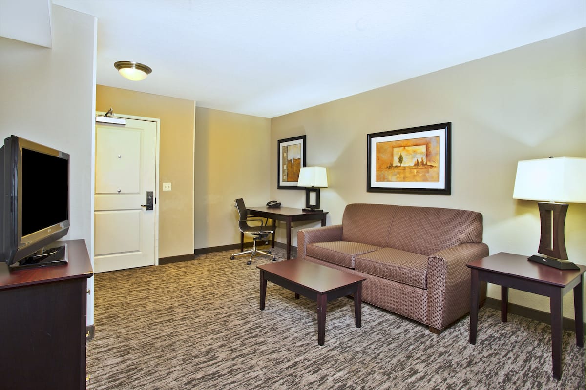 A hotel room with a sofa, a coffee table, a TV on a stand, a desk with a chair, and framed artwork on the wall.