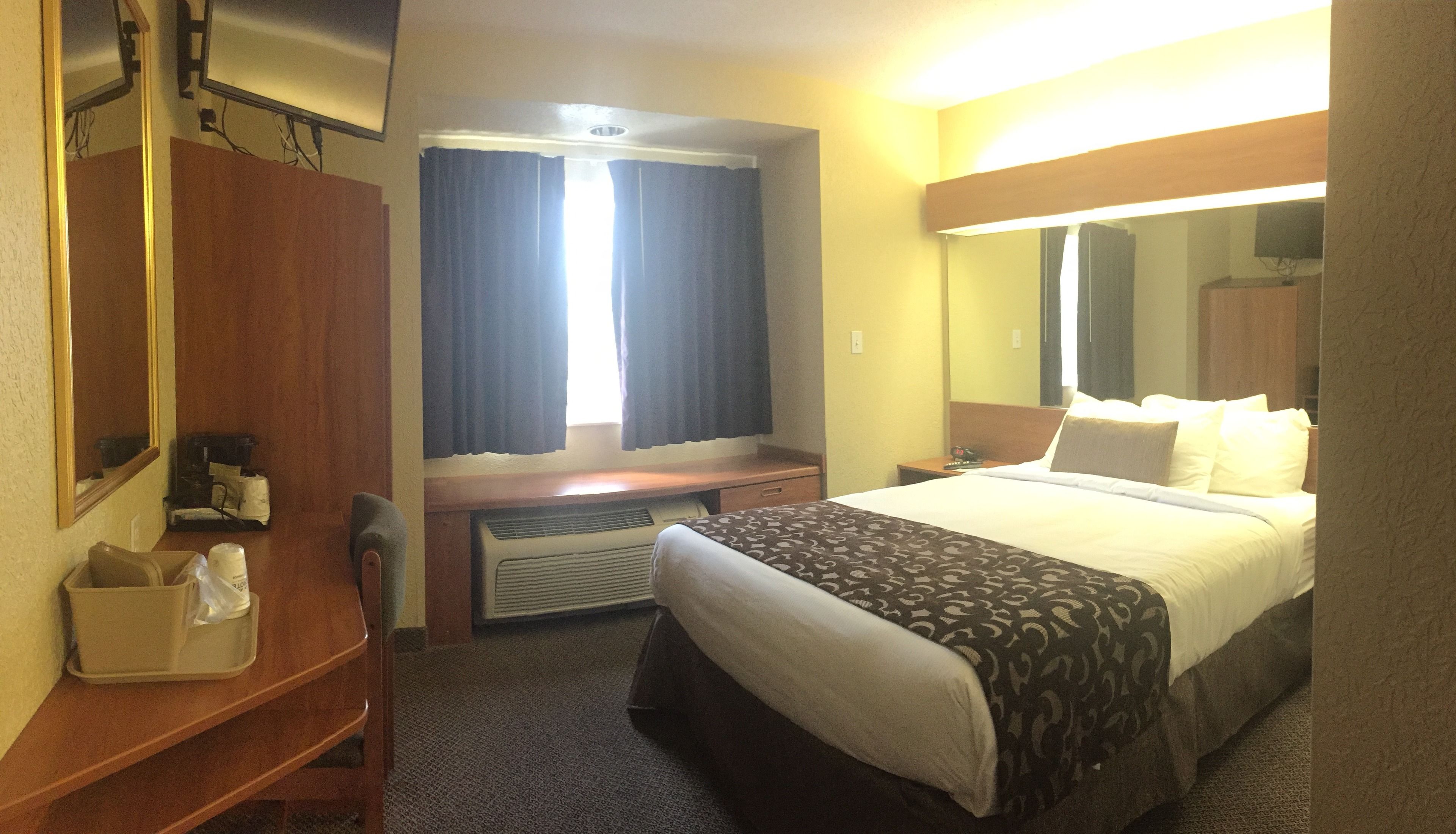 A hotel room with a bed, a desk, a chair, a mirror, a window with curtains, and a wall-mounted TV.