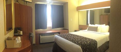 A hotel room with a bed, a desk, a chair, a mirror, a window with curtains, and a wall-mounted TV.