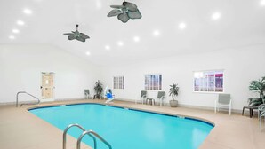 Una piscina interna con acqua cristallina blu, circondata da pareti bianche e un pavimento piastrellato. Sono presenti ventilatori a soffitto, una porta e una finestra con vista.