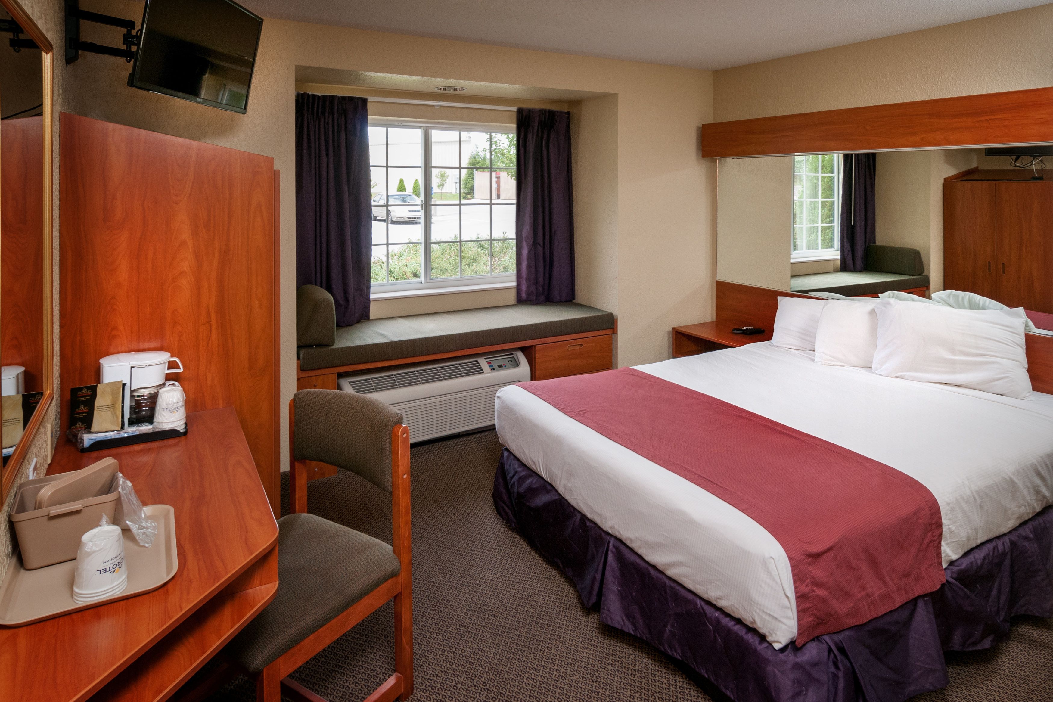 A hotel room with a large bed, a desk with a chair, a window with curtains, a television mounted on the wall, and a small table with a tray.