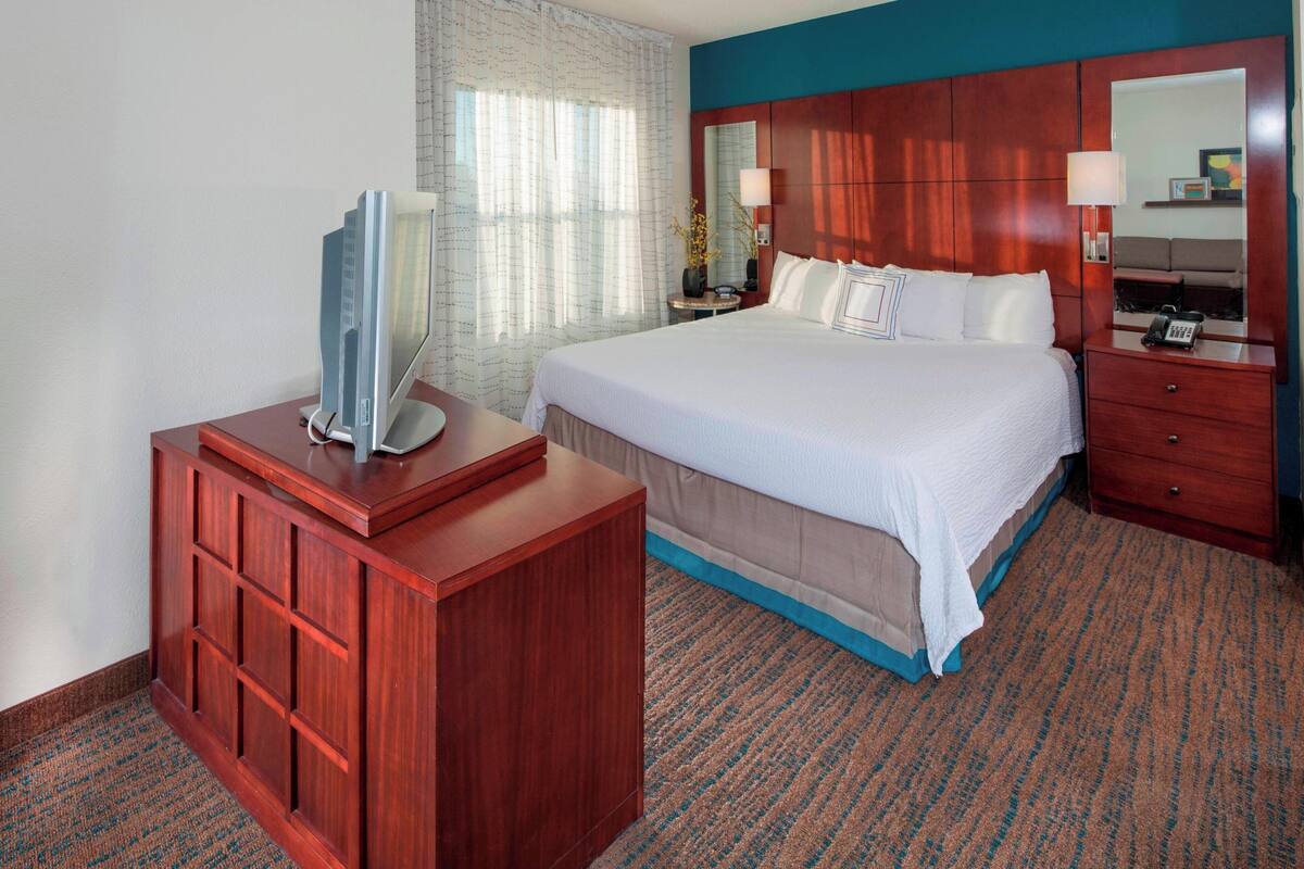 Habitación de hotel con una cama grande, un aparador de madera, un televisor y una mesita de noche con lámpara.