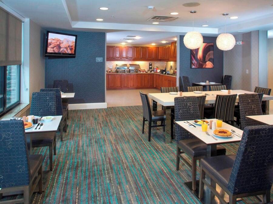 A hotel dining area with a view of the kitchen, tables with chairs, and a television on the wall.