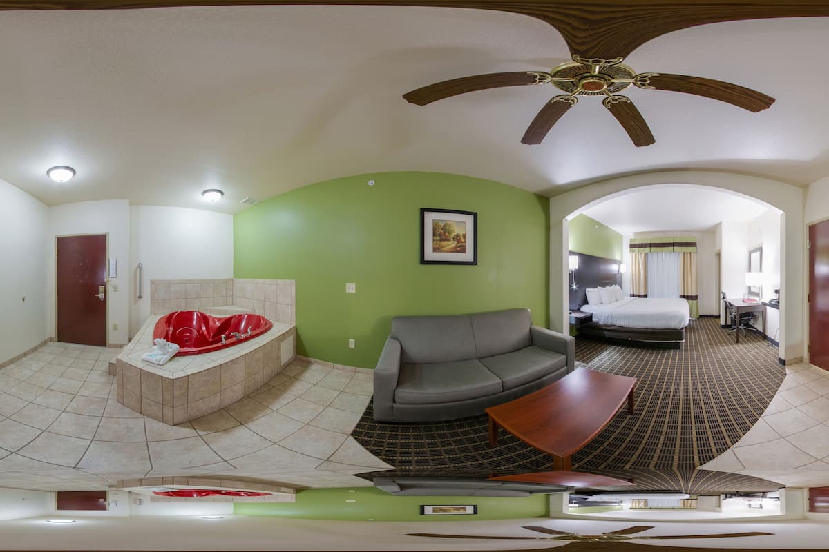 A panoramic view of a hotel room with a curved bathtub, a gray sofa, a bed, a TV, and a ceiling fan.