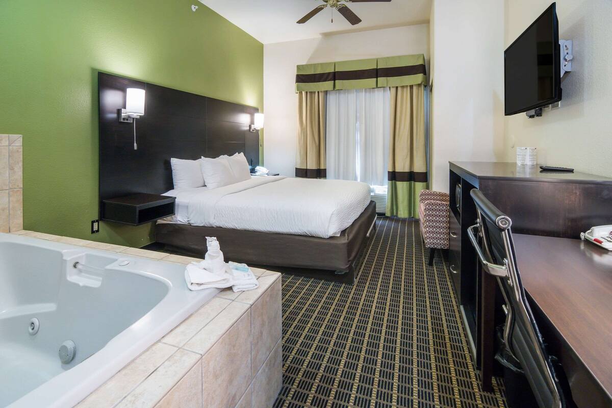 A hotel room with a large bed, a separate bathroom with a bathtub, a desk with a chair, and a flat-screen TV mounted on the wall.