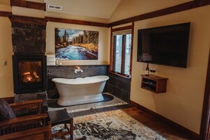 A bathroom with a stone fireplace, a freestanding bathtub, a large window, and a mounted television.