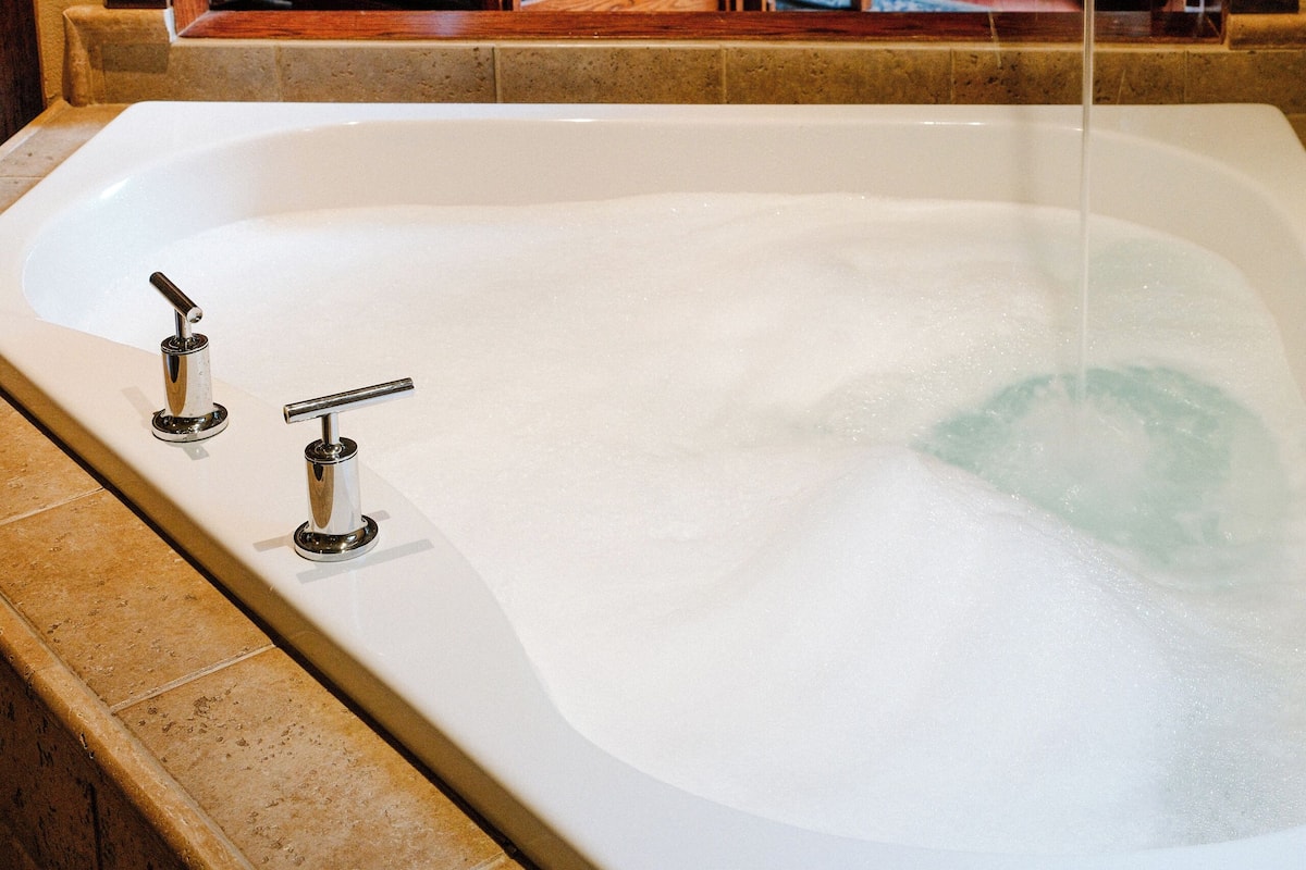 A bathtub with two faucets, a wooden chair, and a fireplace in the background.