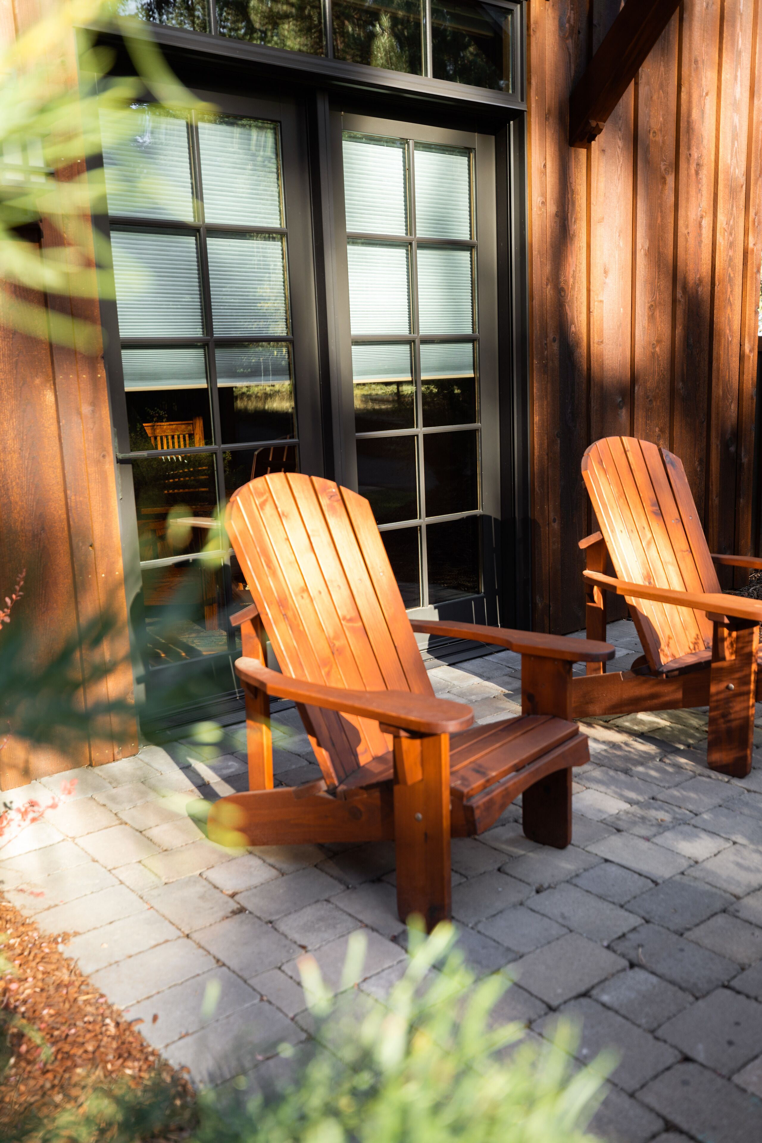Two wooden Adirondack chairs on a paved patio with a wooden fence and a window in the background.