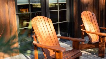 Dos sillas Adirondack de madera en un patio empedrado, con una cerca de madera y una ventana al fondo.