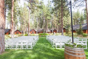 A forested outdoor venue with rows of white chairs arranged for an event, surrounded by tall trees and wooden cabins.