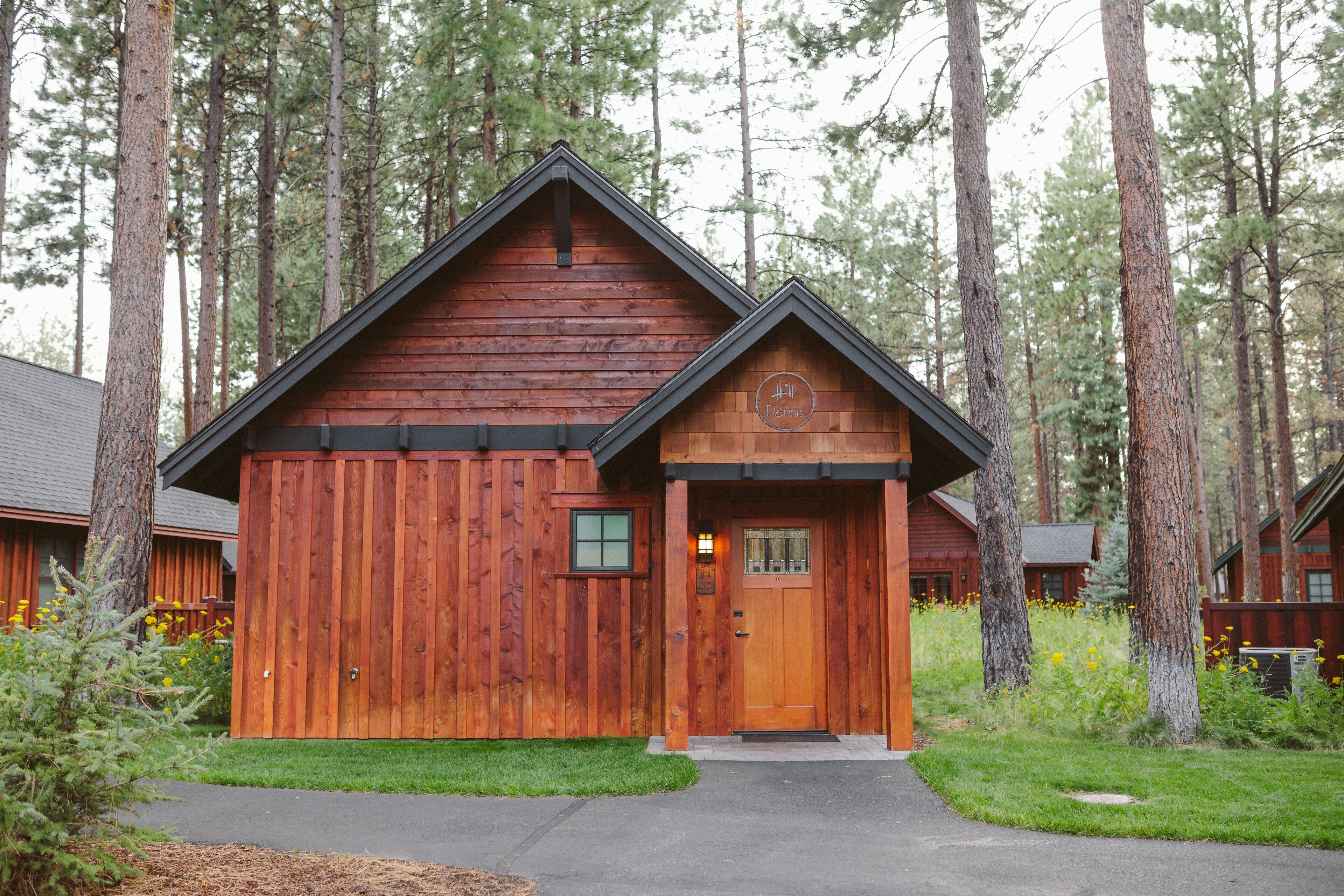 A wooden cabin with a front door and window, surrounded by tall trees and a well-maintained lawn.