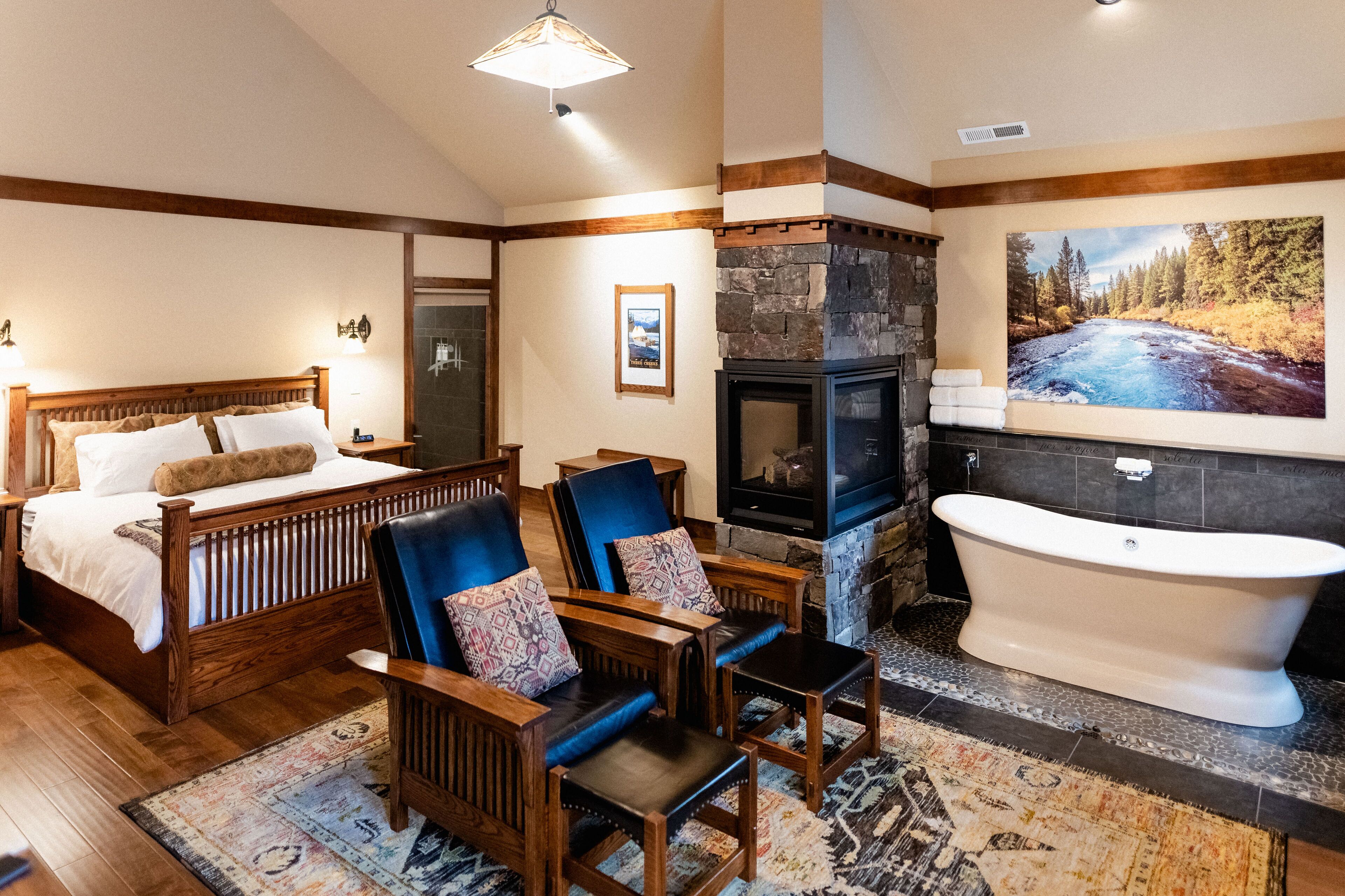 Een slaapkamer met een houten bed, twee fauteuils, een stenen openhaard, een groot bad en een schilderij van een rivier aan de muur.
