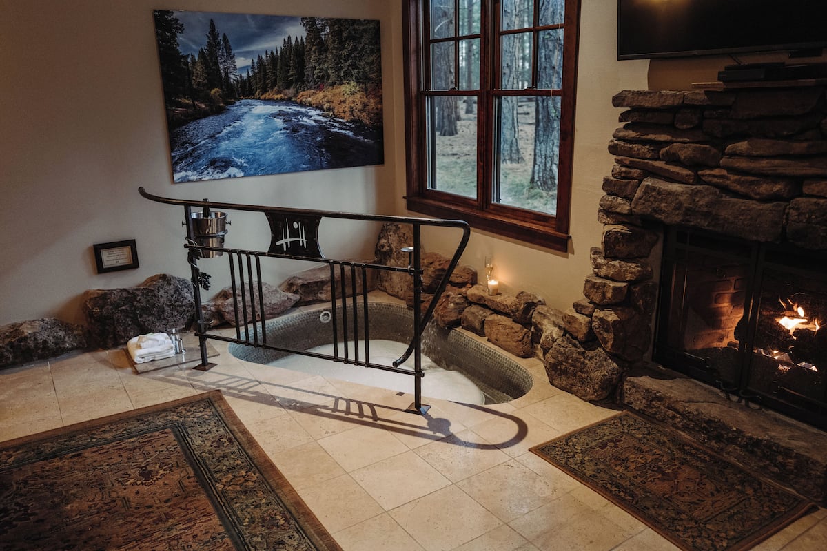 A cozy room with a stone fireplace, a large painting of a river, and a window with a view of trees.