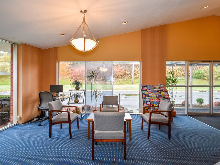 Un hall d’entrée spacieux, doté d’une moquette bleue, de chaises en bois, d’une grande fenêtre et d’une bibliothèque.