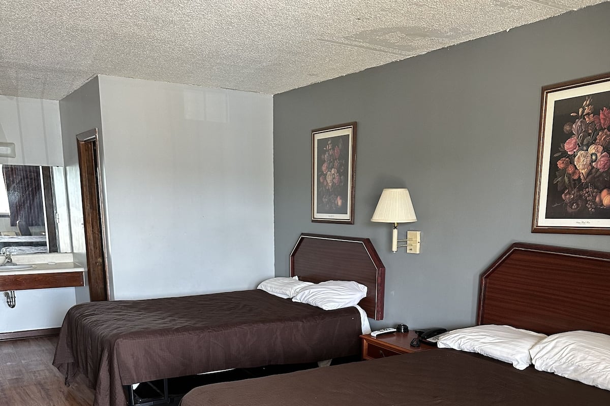 A hotel room with two beds, a wooden floor, a desk with a lamp, and a painting on the wall.