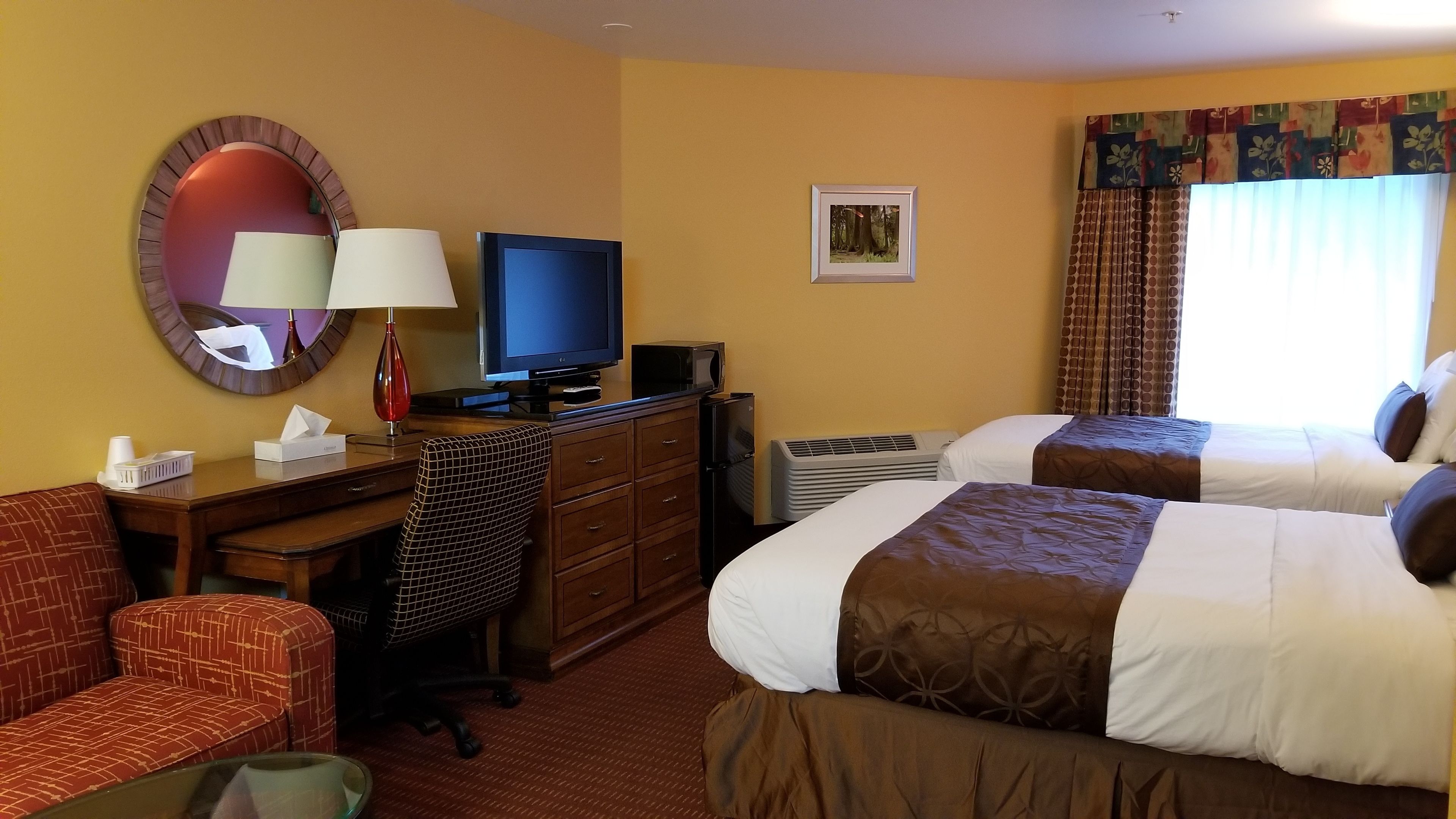 A hotel room with two beds, a desk with a computer, a TV, a chair, a couch, and a window with curtains.