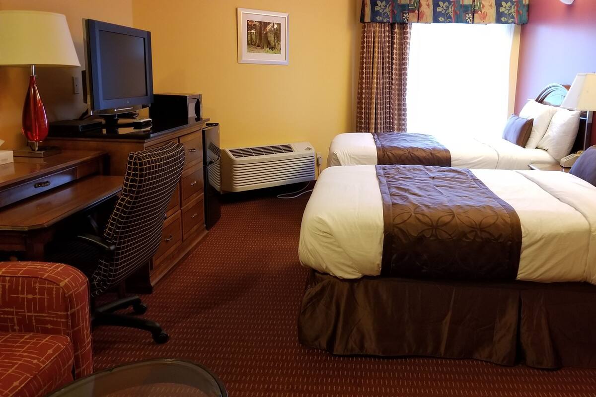 A hotel room with two beds, a desk with a television and a lamp, a chair, and an air conditioning unit.