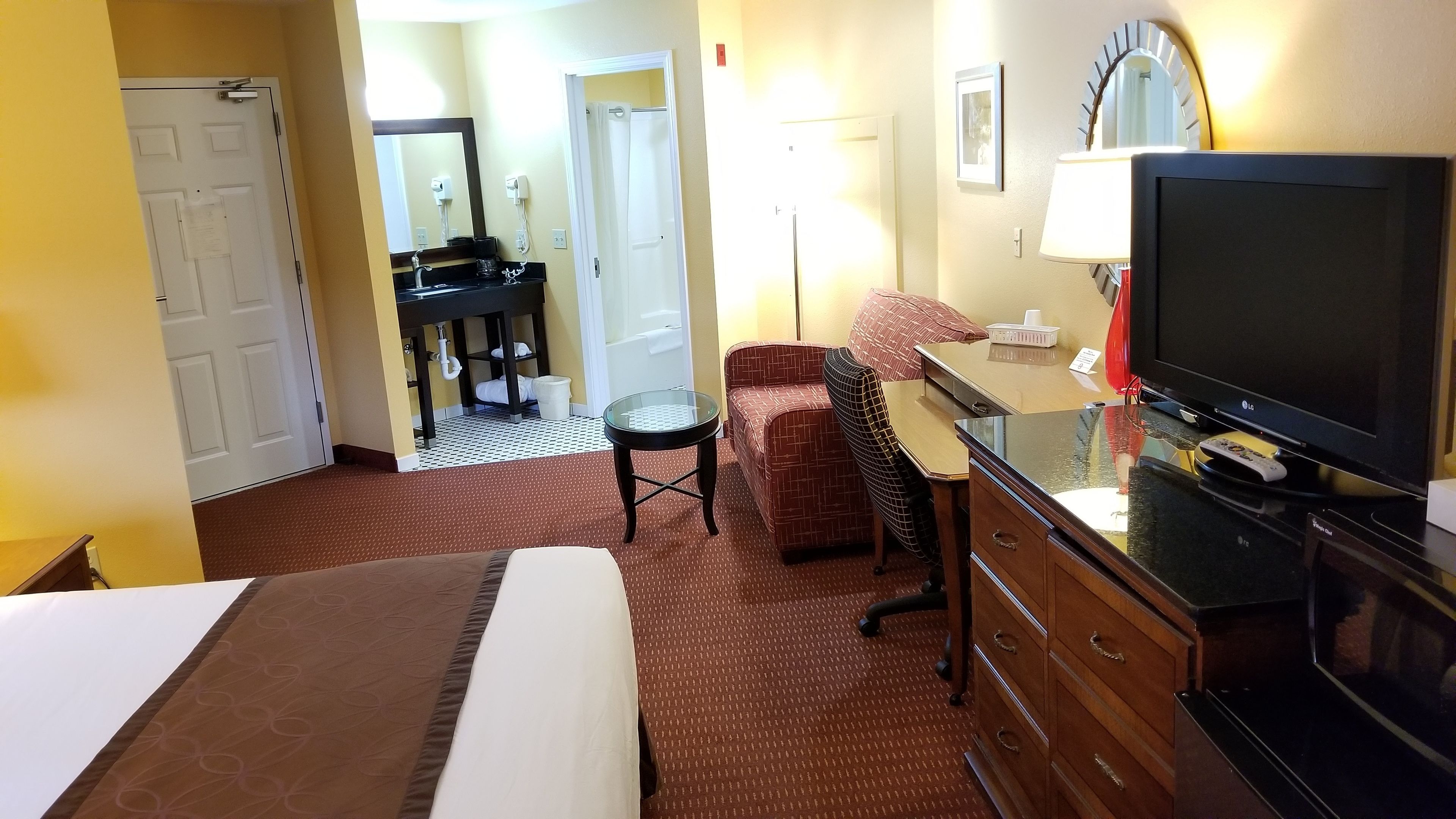 Quarto de hotel com cama, televisão, secretária com abajur, cadeira e banheiro visível por uma porta aberta.