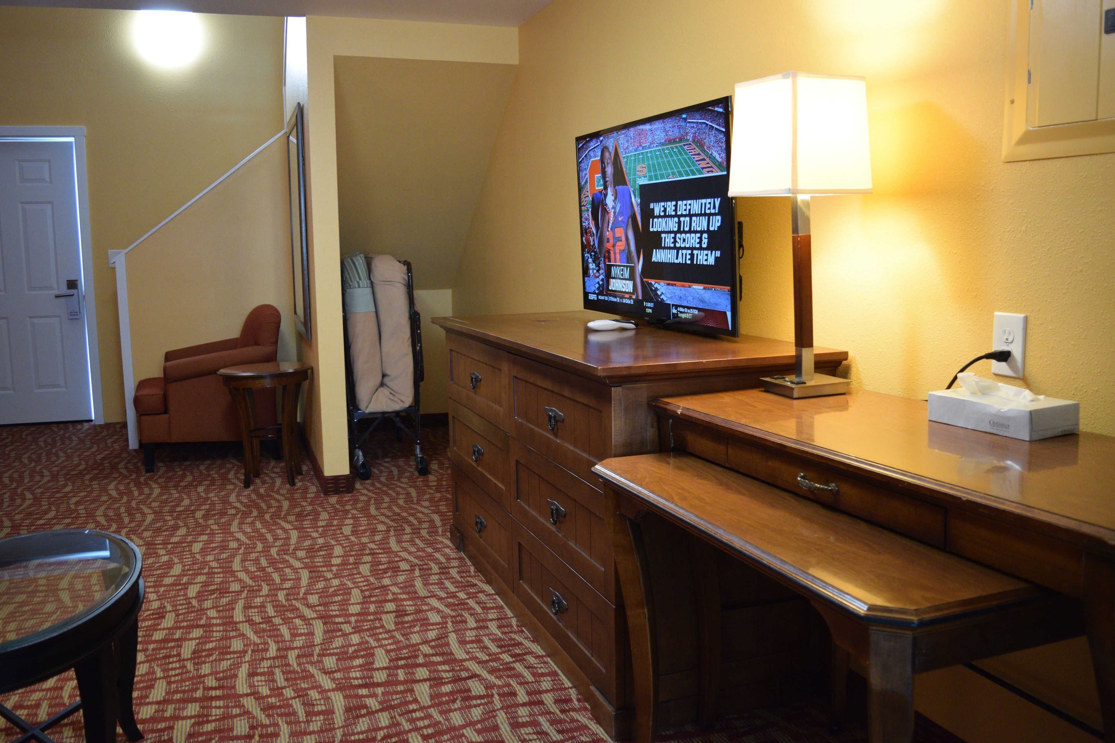 A hotel room with a wooden desk, a television, a lamp, a chair, and a coat rack.