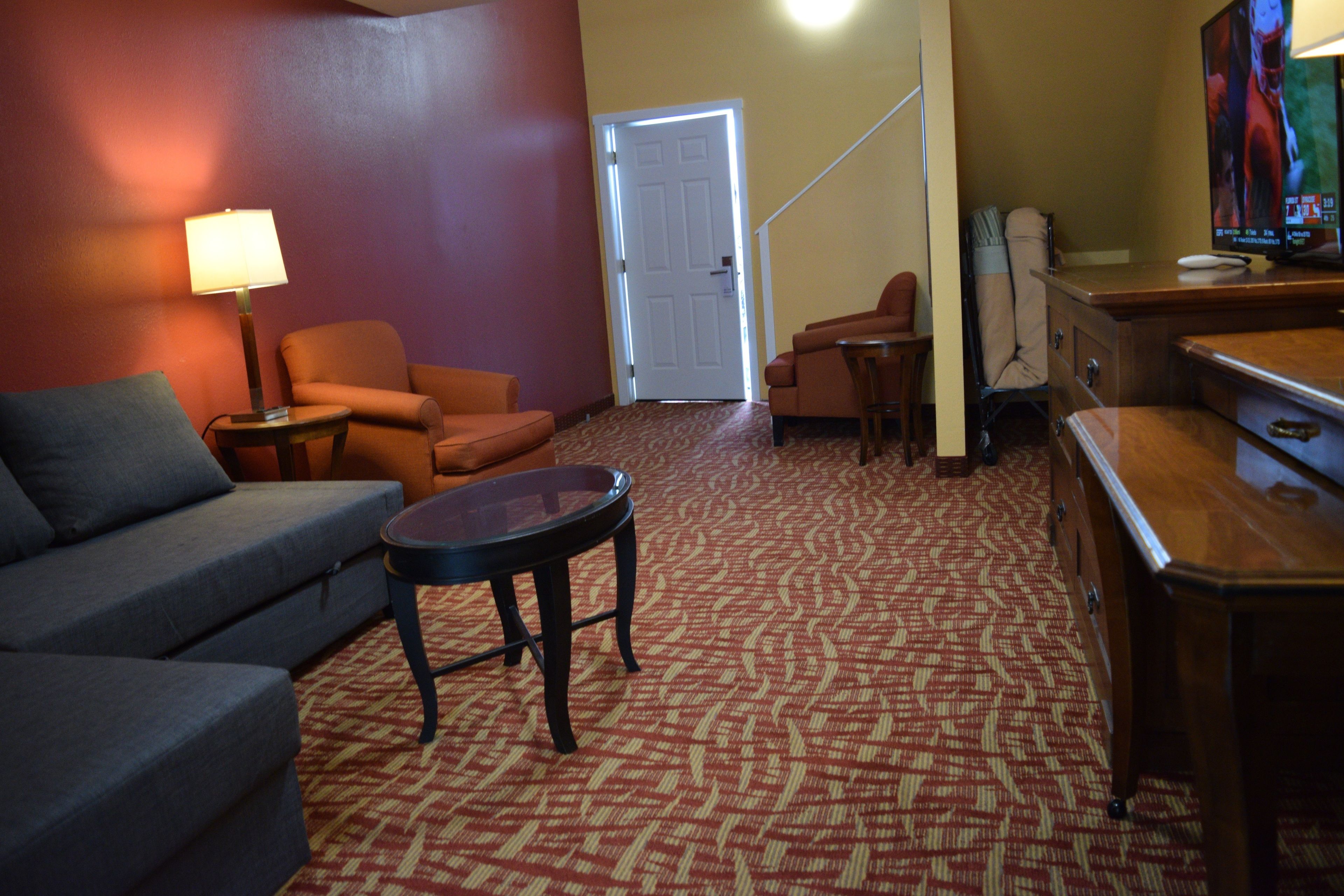 A living room with a sofa, armchair, and a small round table. There is a door leading to another room and a television on a stand.