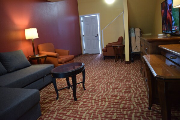 A living room with a sofa, armchair, and a small round table. There is a door leading to another room and a television on a stand.