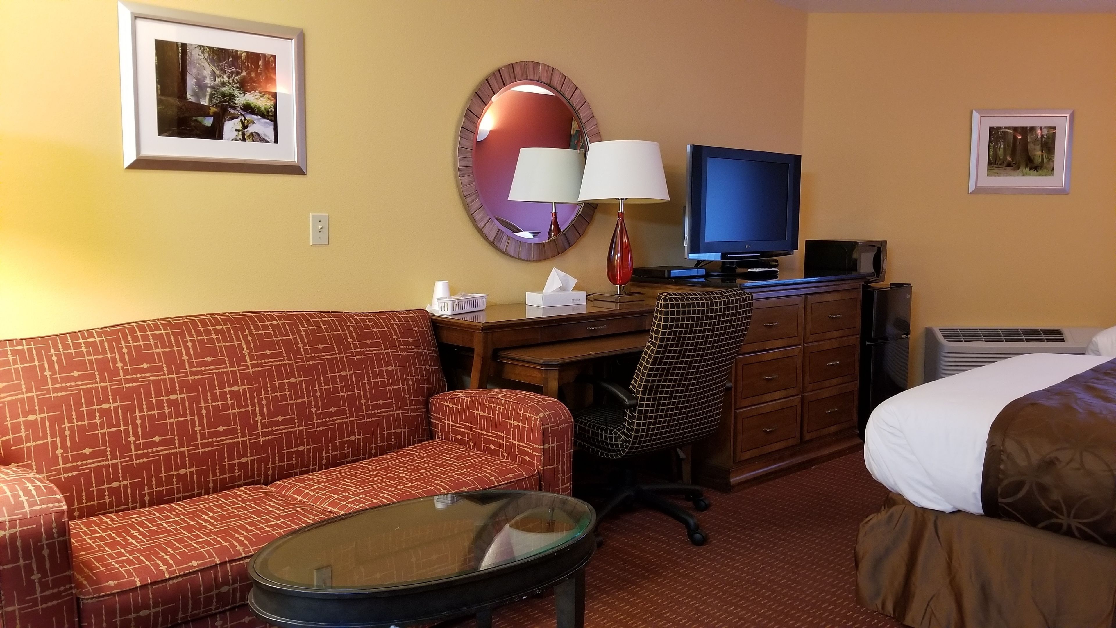 A hotel room with a red patterned sofa, a glass coffee table, a desk with a computer and lamp, a television, and a bed with white and brown bedding.