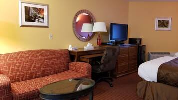 A hotel room with a red patterned sofa, a glass coffee table, a desk with a computer and lamp, a television, and a bed with white and brown bedding.