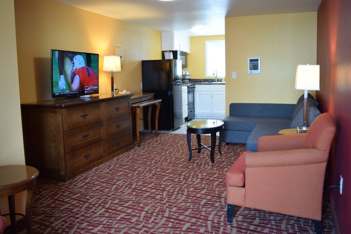 A hotel room with a TV, wooden dresser, sofa, and a kitchenette.