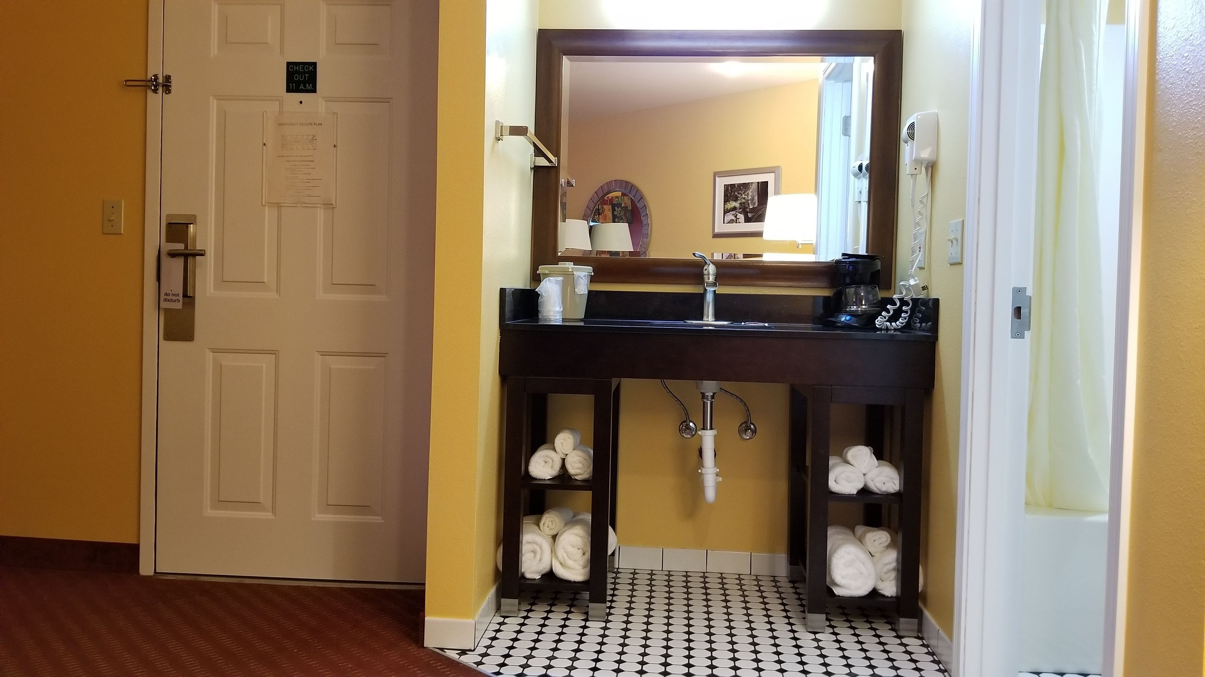 A bathroom with a sink, mirror, towel racks, and a door with a 'CHECK OUT' sign.