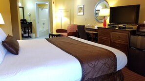 A hotel room with a large bed, a desk, a chair, a television, and a refrigerator.