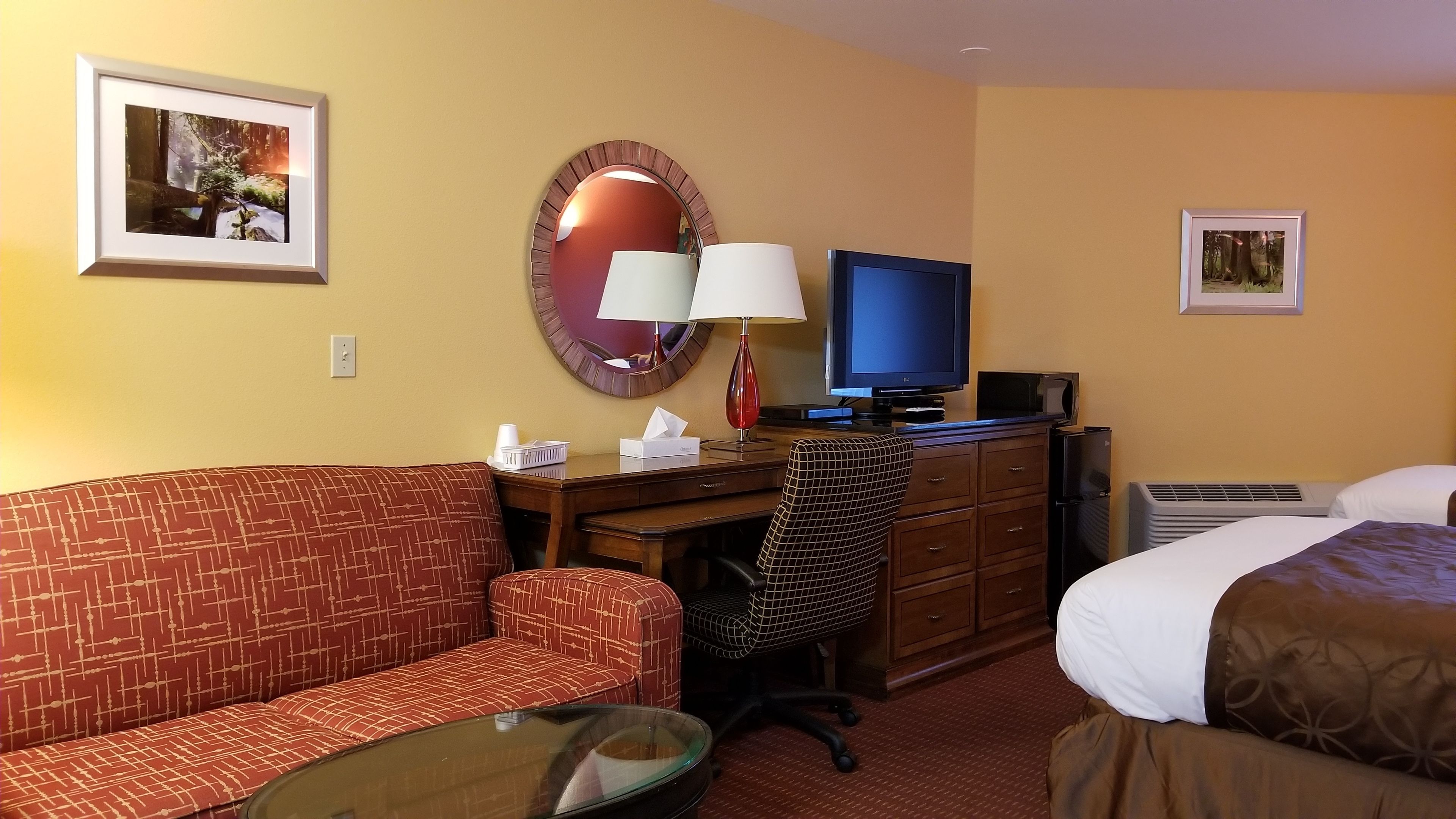 A hotel room with a red patterned sofa, a desk with a lamp, a television, a dresser, and a bed with a brown blanket.