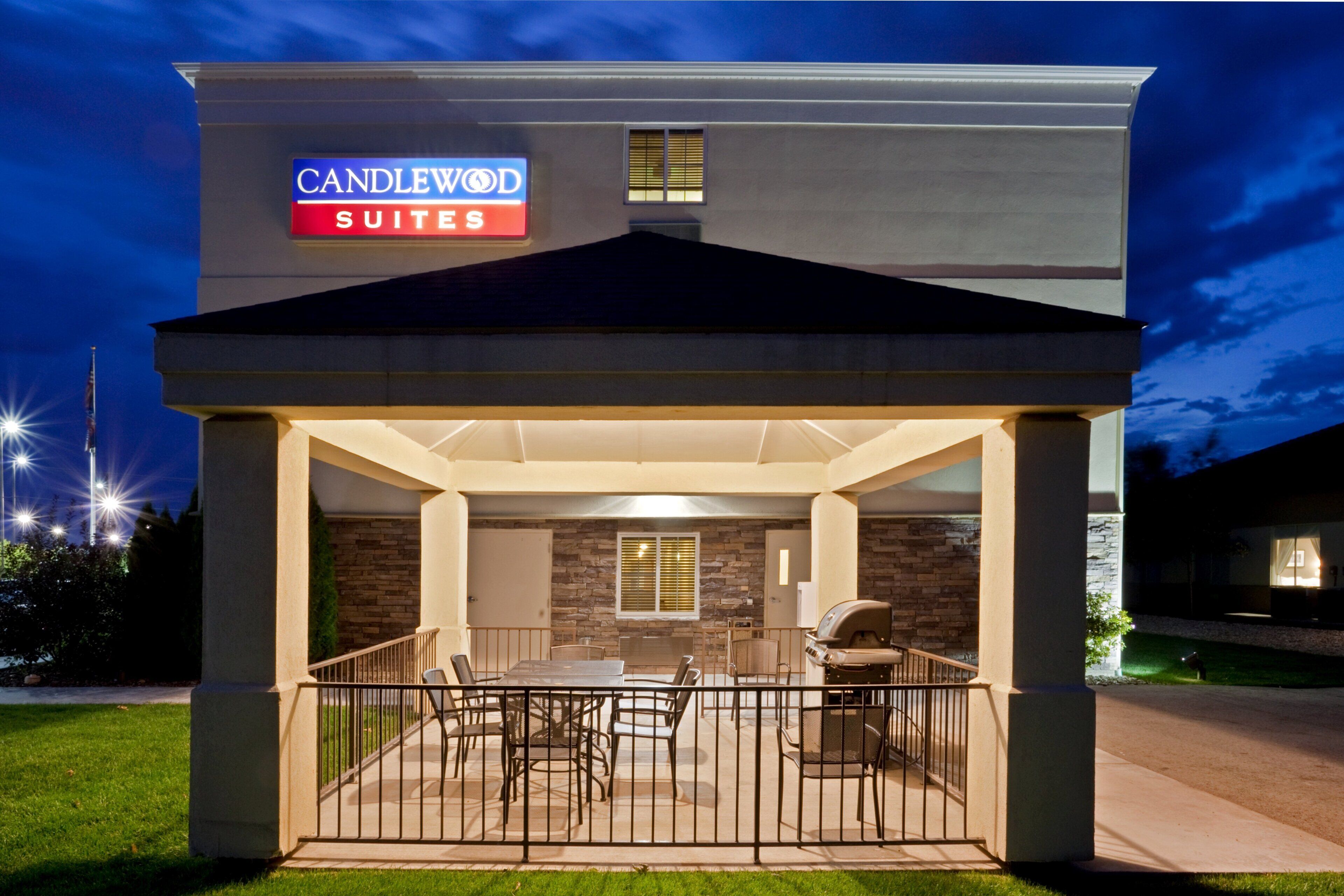 Exterior de un hotel con un patio cubierto, mobiliario exterior y un cartel que indica 'CANDLEWOOD SUITES'.