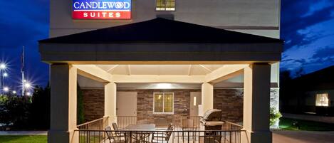 Exterior de un hotel con un patio cubierto, mobiliario exterior y un cartel que indica 'CANDLEWOOD SUITES'.