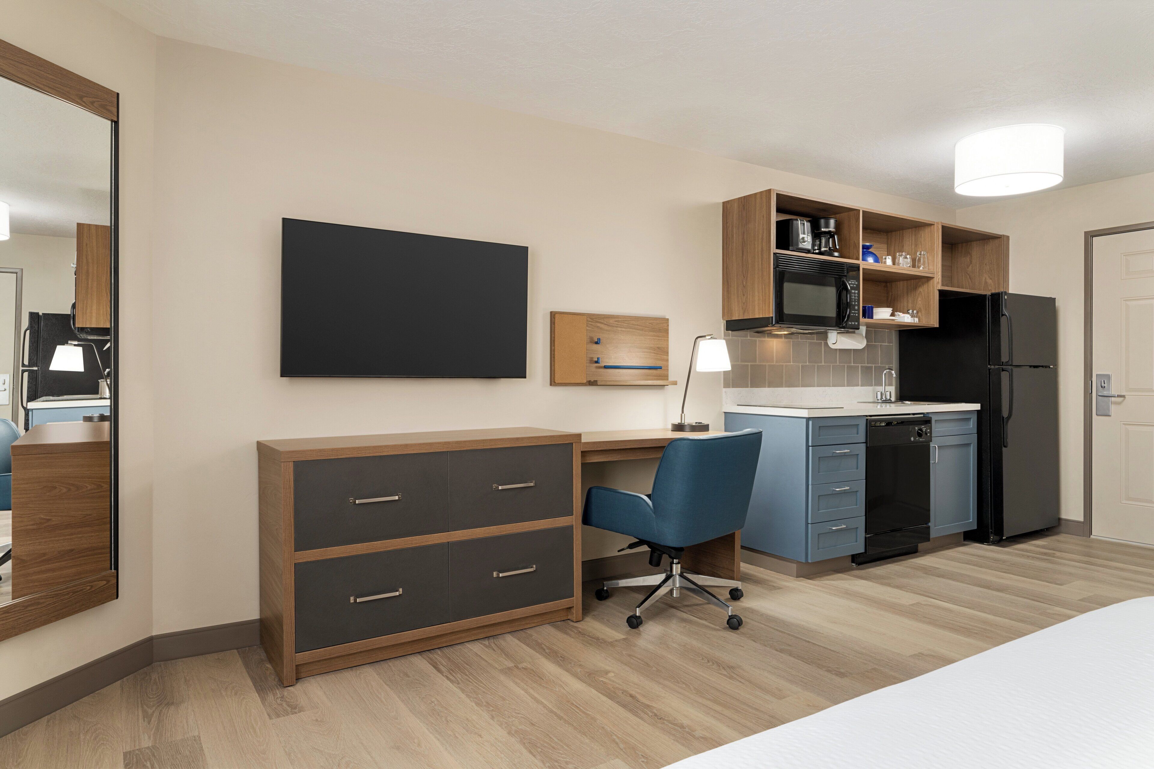 Une chambre d’hôtel moderne dotée d’un bureau en bois, d’un fauteuil de bureau bleu, d’un téléviseur à écran plat, d’un four à micro-ondes encastré, d’un réfrigérateur et d’une petite kitchenette.