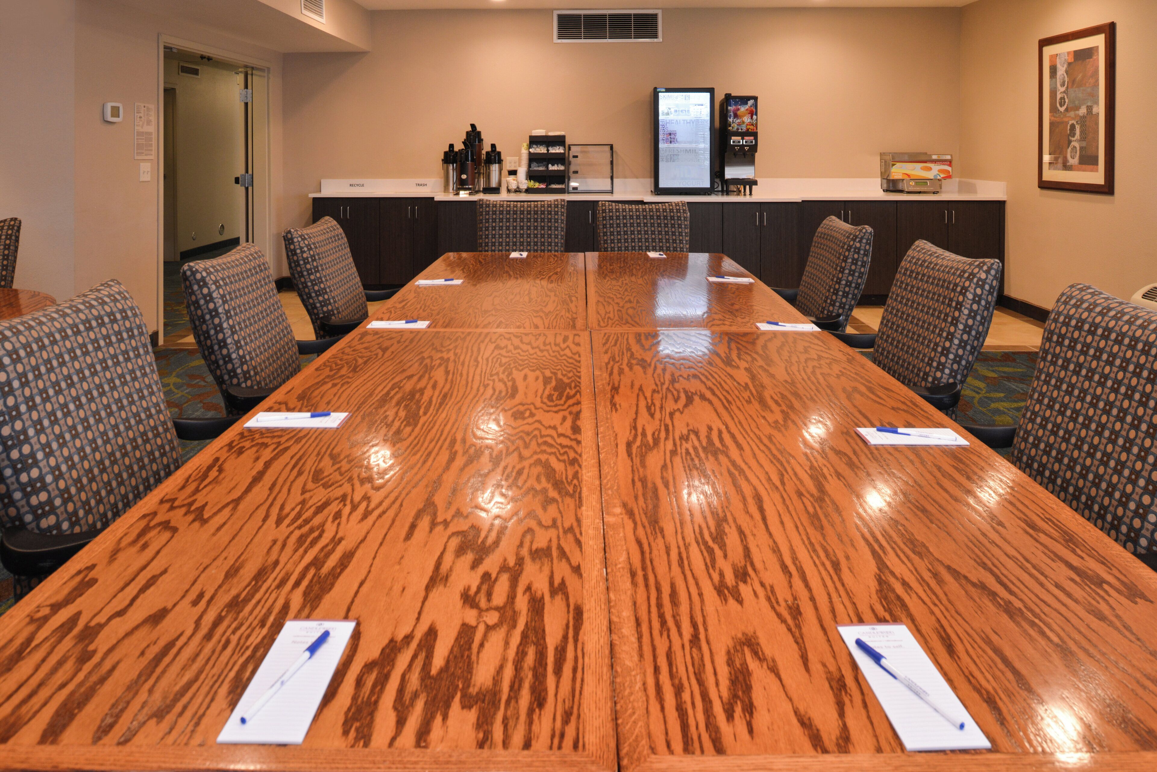 Un salón de conferencias con una gran mesa de madera, seis sillas estampadas, una máquina de café y un refrigerador.
