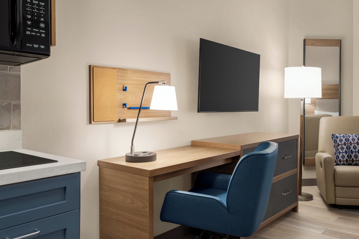 Una moderna camera d'albergo con una scrivania in legno, una poltrona blu, una televisione a schermo piatto, una lampada, un divano con cuscini decorati e uno specchio.