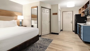 A hotel room with a bed, bedside lamp, a mirror, a door, a kitchen area with a refrigerator, microwave, and cabinets. - Candlewood Suites Boise - Towne Square by IHG (Boise)