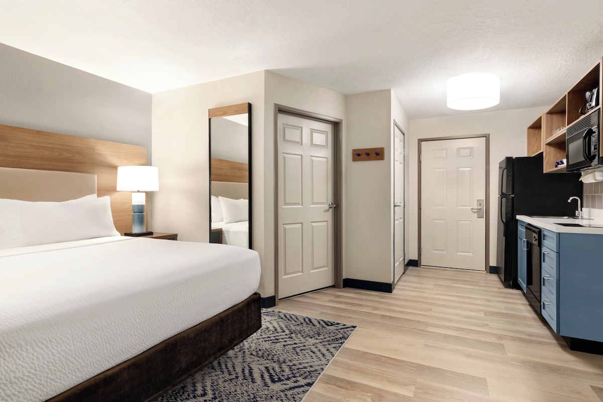 A hotel room with a bed, bedside lamp, a mirror, a door, a kitchen area with a refrigerator, microwave, and cabinets.