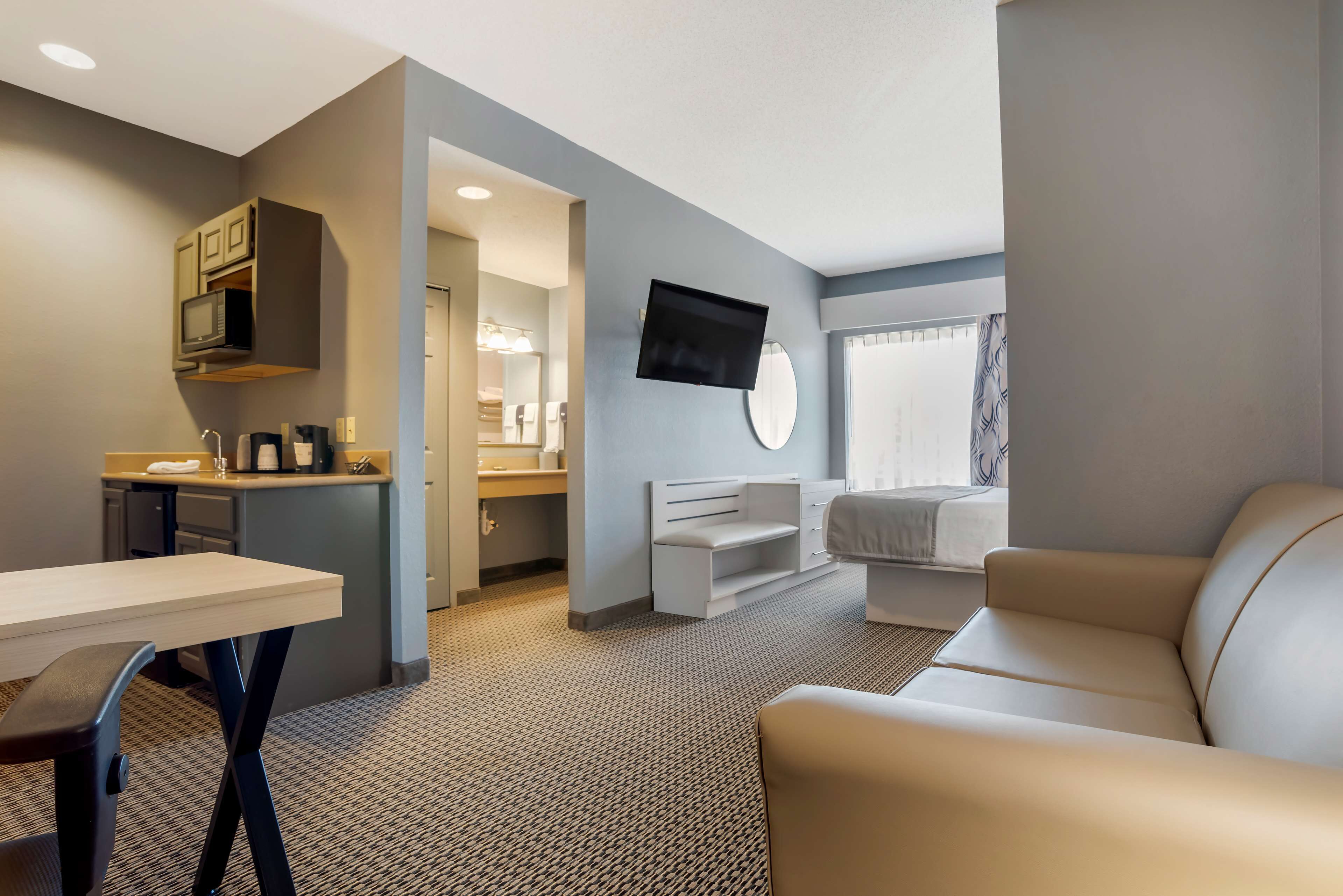 A modern hotel room with a kitchenette, a flat-screen TV, a sofa, and a dining area.