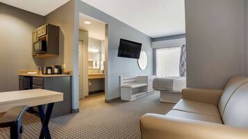 A modern hotel room with a kitchenette, a flat-screen TV, a sofa, and a dining area.