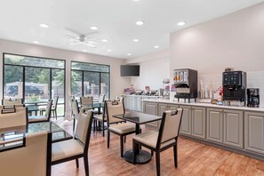 A modern dining area with glass-top tables and chairs, a kitchen with a coffee machine, and large windows offering a view of the outdoors.