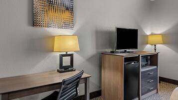 A hotel room with a wooden desk, a chair, a television, a minibar, and two lamps.