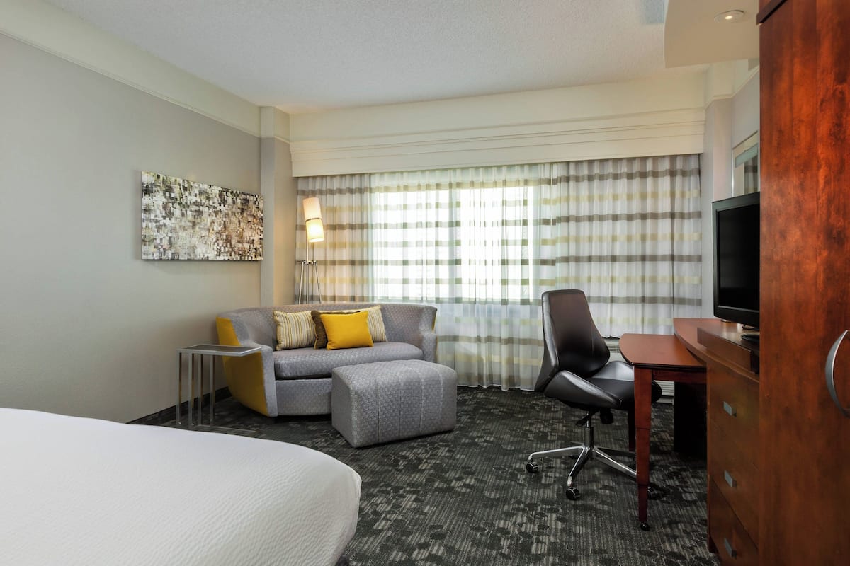Une chambre d’hôtel comprenant un lit, un canapé, un pouf, un bureau, une télévision et une grande fenêtre avec des rideaux.