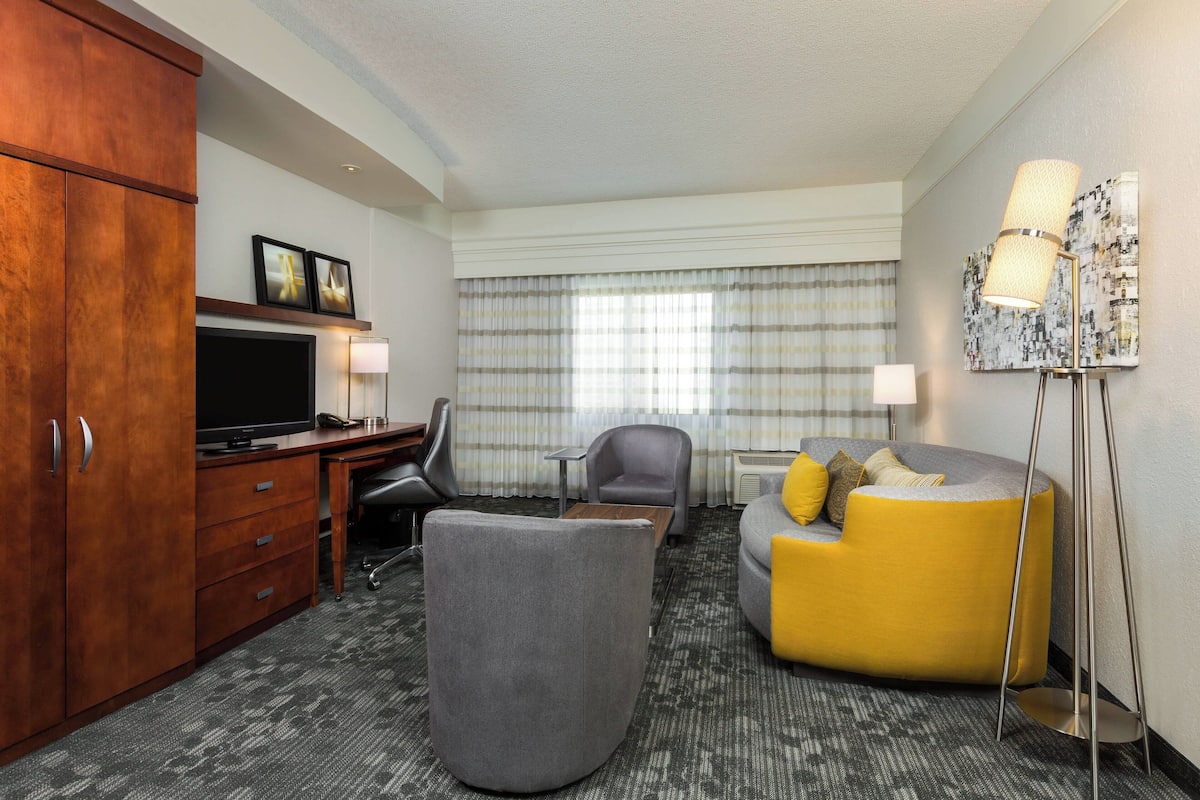 Une chambre d’hôtel avec une armoire en bois, un téléviseur à écran plat, un canapé gris, un canapé jaune, un fauteuil gris, une petite table et un lampadaire.