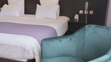 Quarto de hotel com uma cama, uma poltrona verde-água e uma mesa de cabeceira com telefone e abajur.