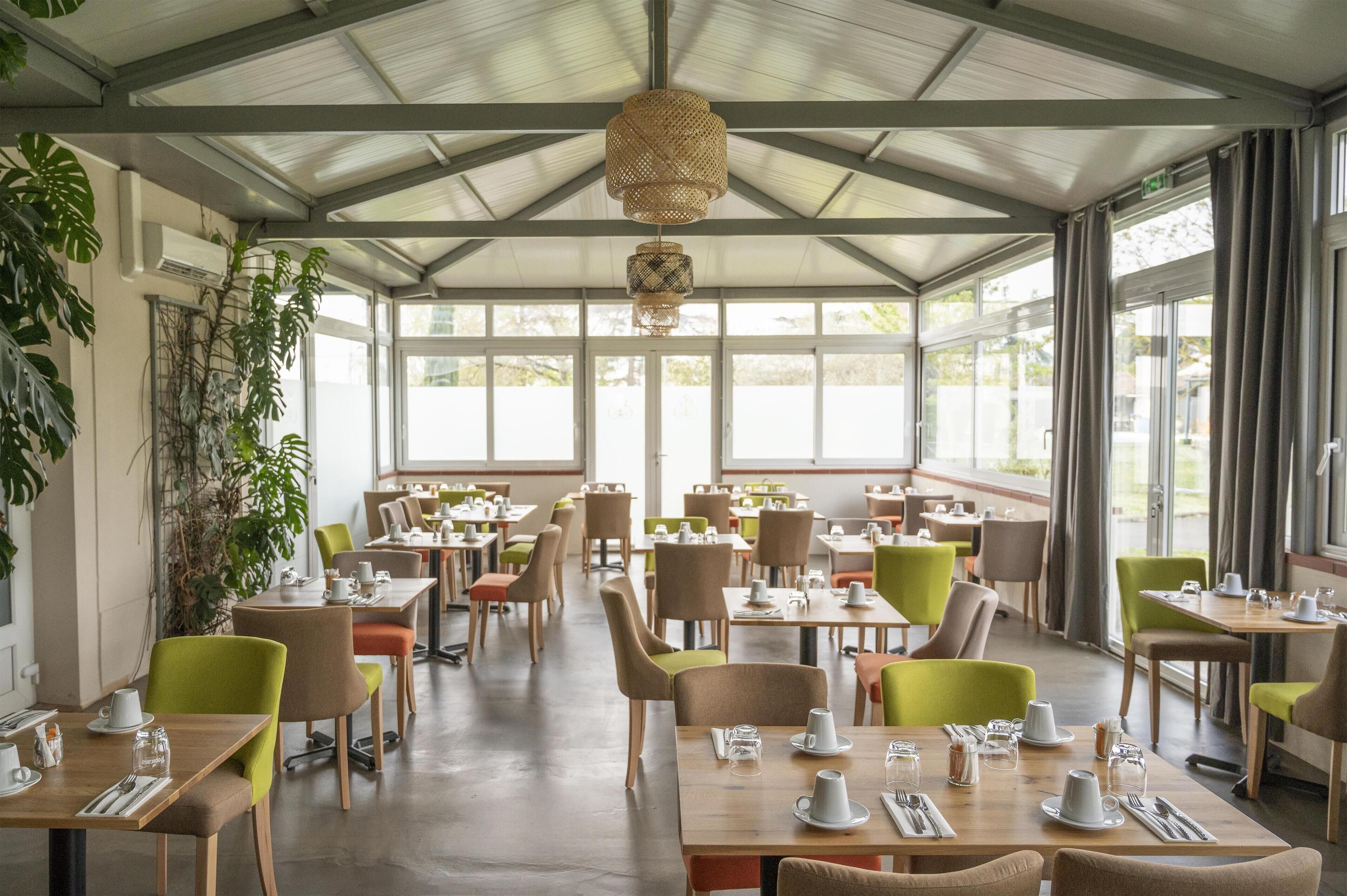 A spacious indoor dining area with multiple tables and chairs, large windows, and hanging light fixtures.