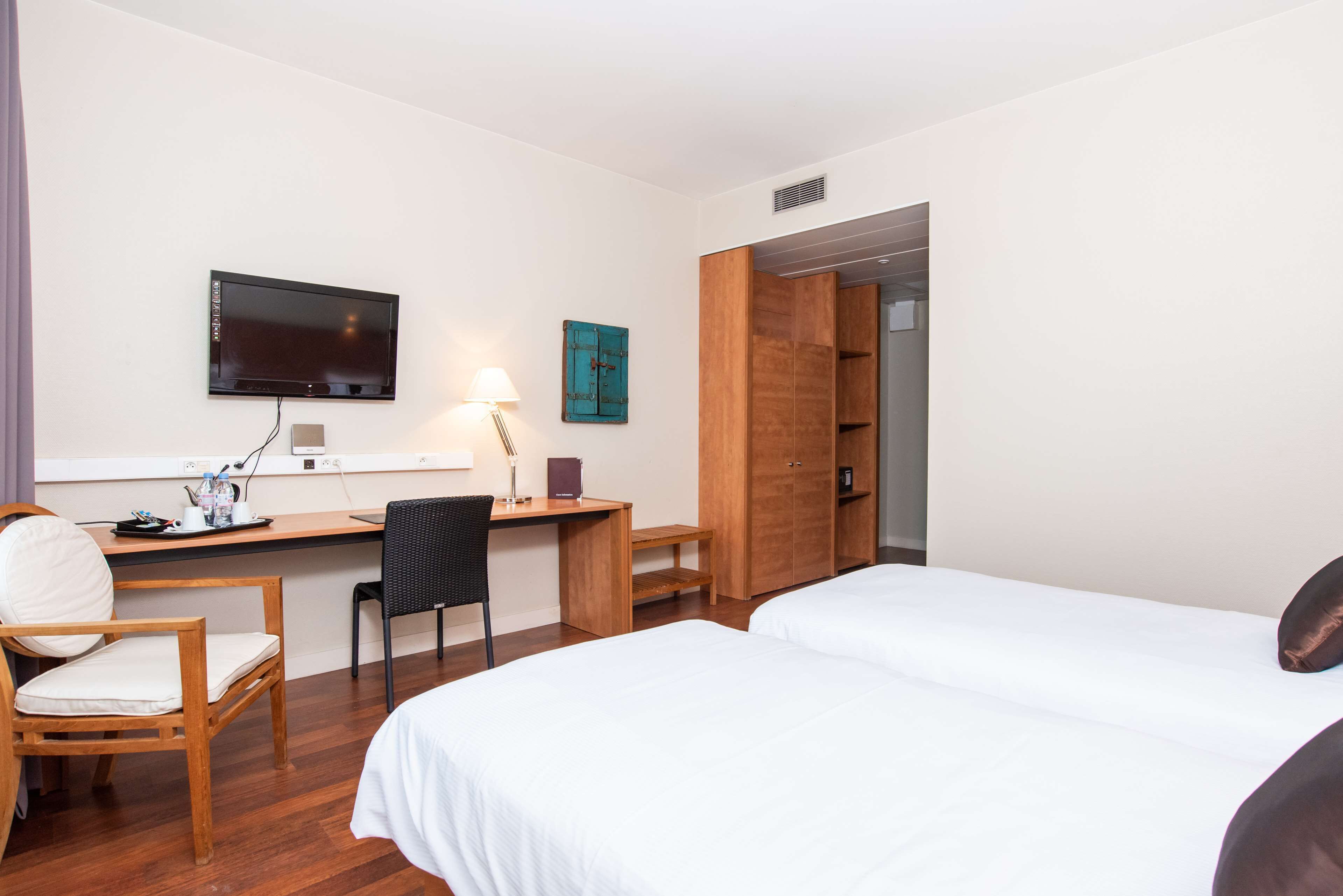 A hotel room with two beds, a desk with a chair, a TV mounted on the wall, and a wooden wardrobe.