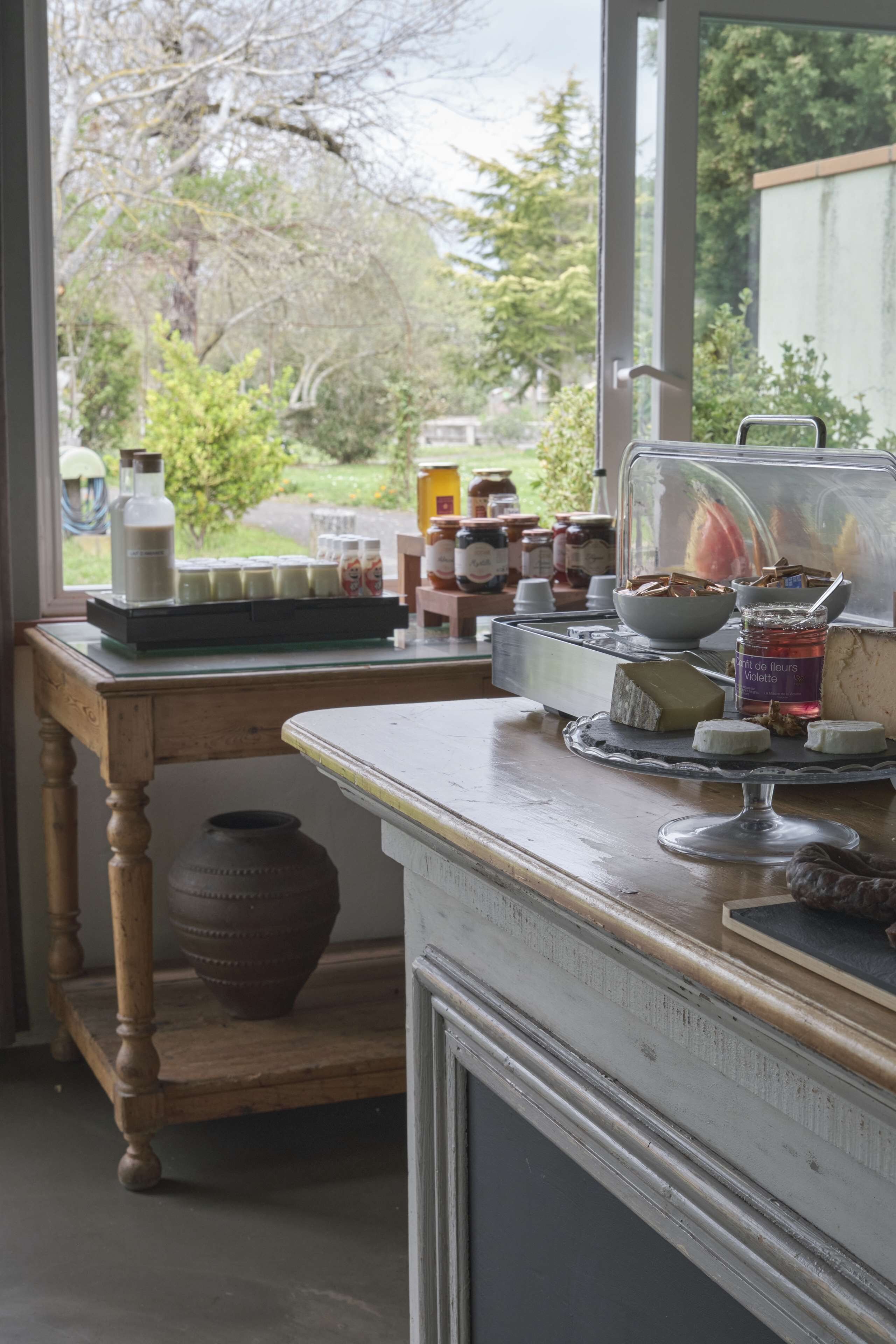 A kitchen counter with various food items and containers, a wooden table with jars, and a view of trees outside.