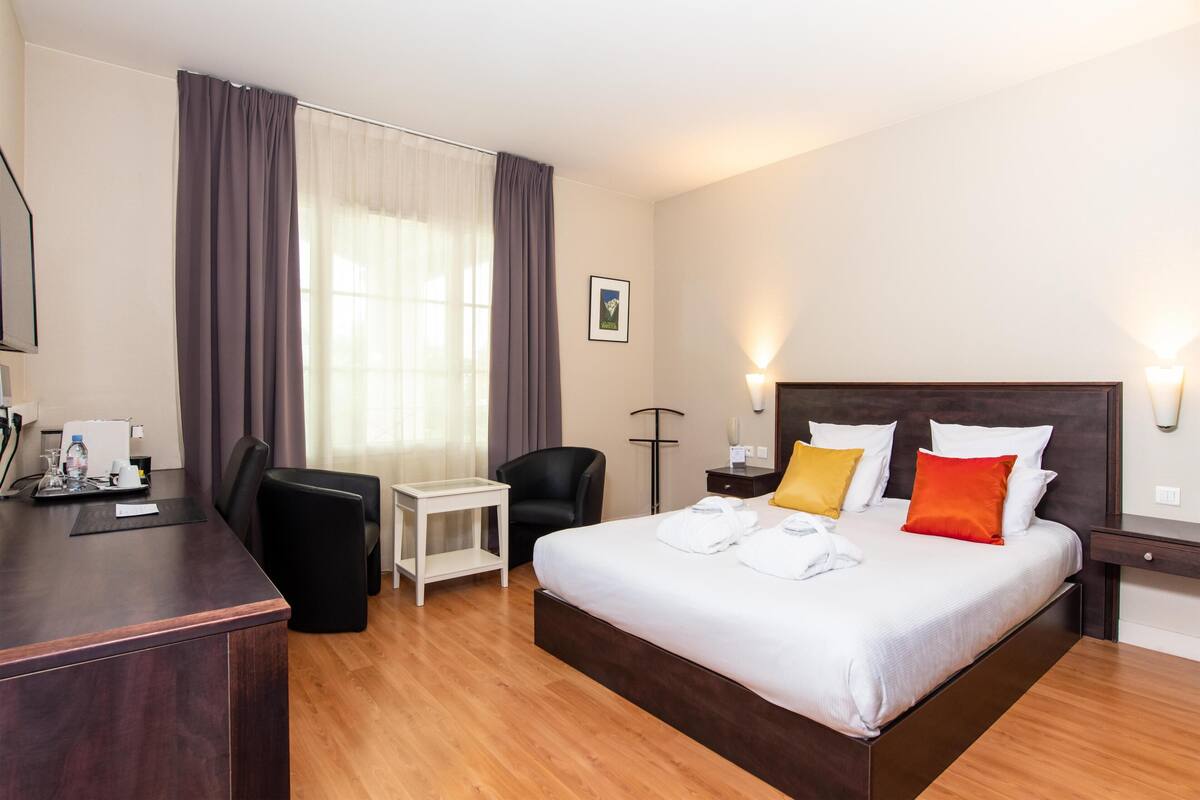 A hotel room with a bed, a desk, a chair, a small table, and a nightstand.